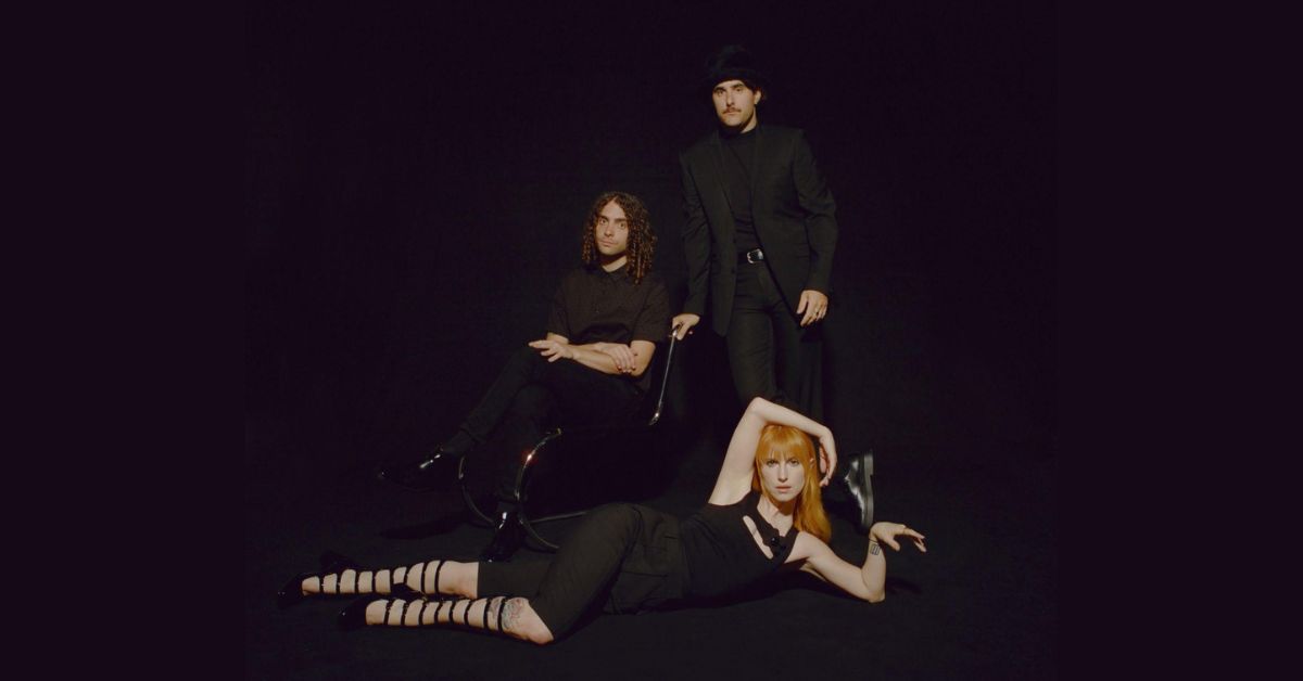 A photo of Paramore against a black wall. Hayley Williams is laying on the ground, across the front of the image.