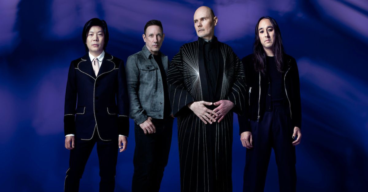 A photo of the Smashing Pumpkins standing against a purple backdrop, singer Billy Corgan is in the front 