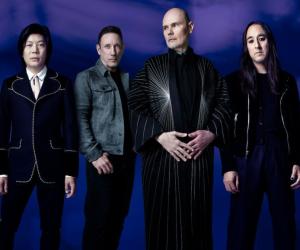 A photo of the Smashing Pumpkins standing against a purple backdrop, singer Billy Corgan is in the front 